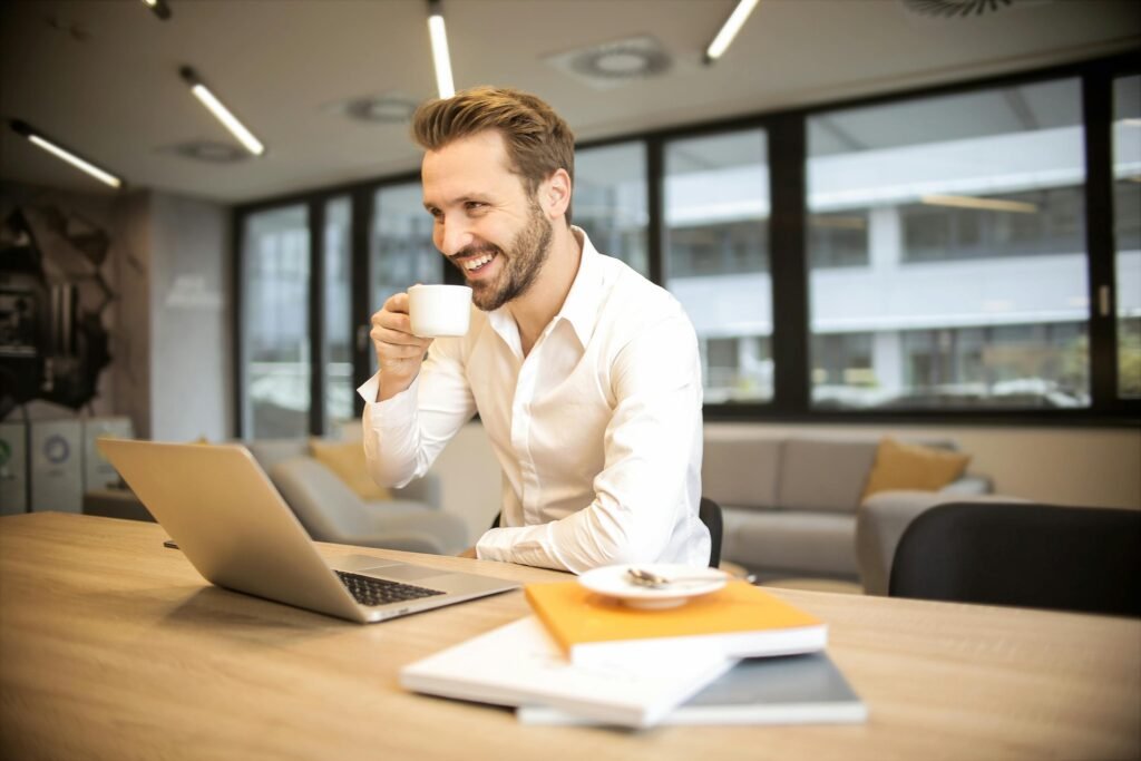 LE GÎTE Man enjoying a coffee break while working in a modern office with laptop and books.